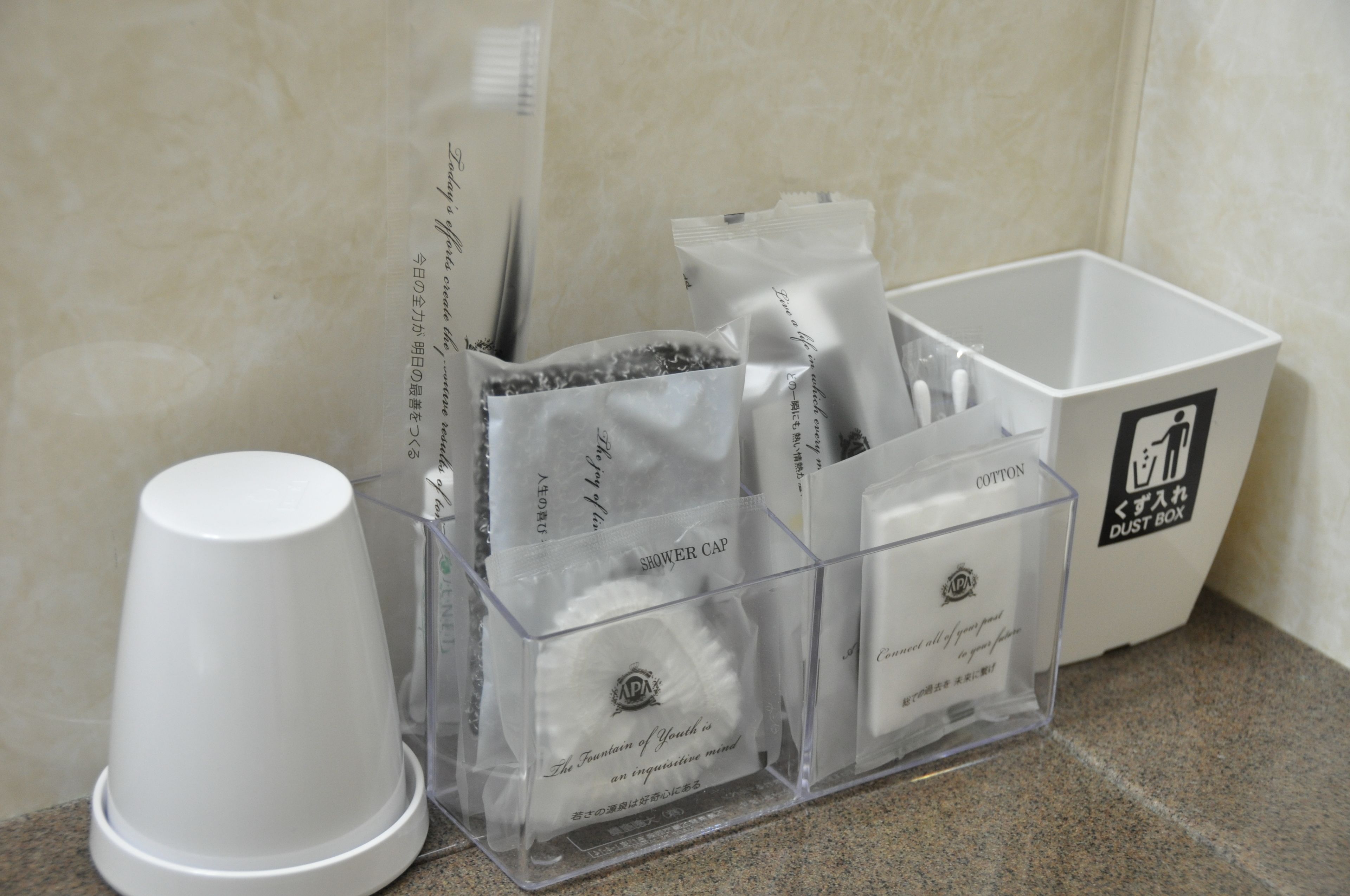 Una encimera de baño con dispensador de gorro de ducha, un bote de algodón y una taza blanca.