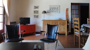 A living room with a TV, two chairs, a wooden cabinet, and a table with remotes. - Villa Lagoon by Agence Bru - 2433 (Lacanau)