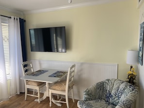 A dining area with a white table and chairs, a flat-screen TV mounted on the wall, and a patterned armchair. - Panoramic sea views in Admirals Row (Hilton Head Island)