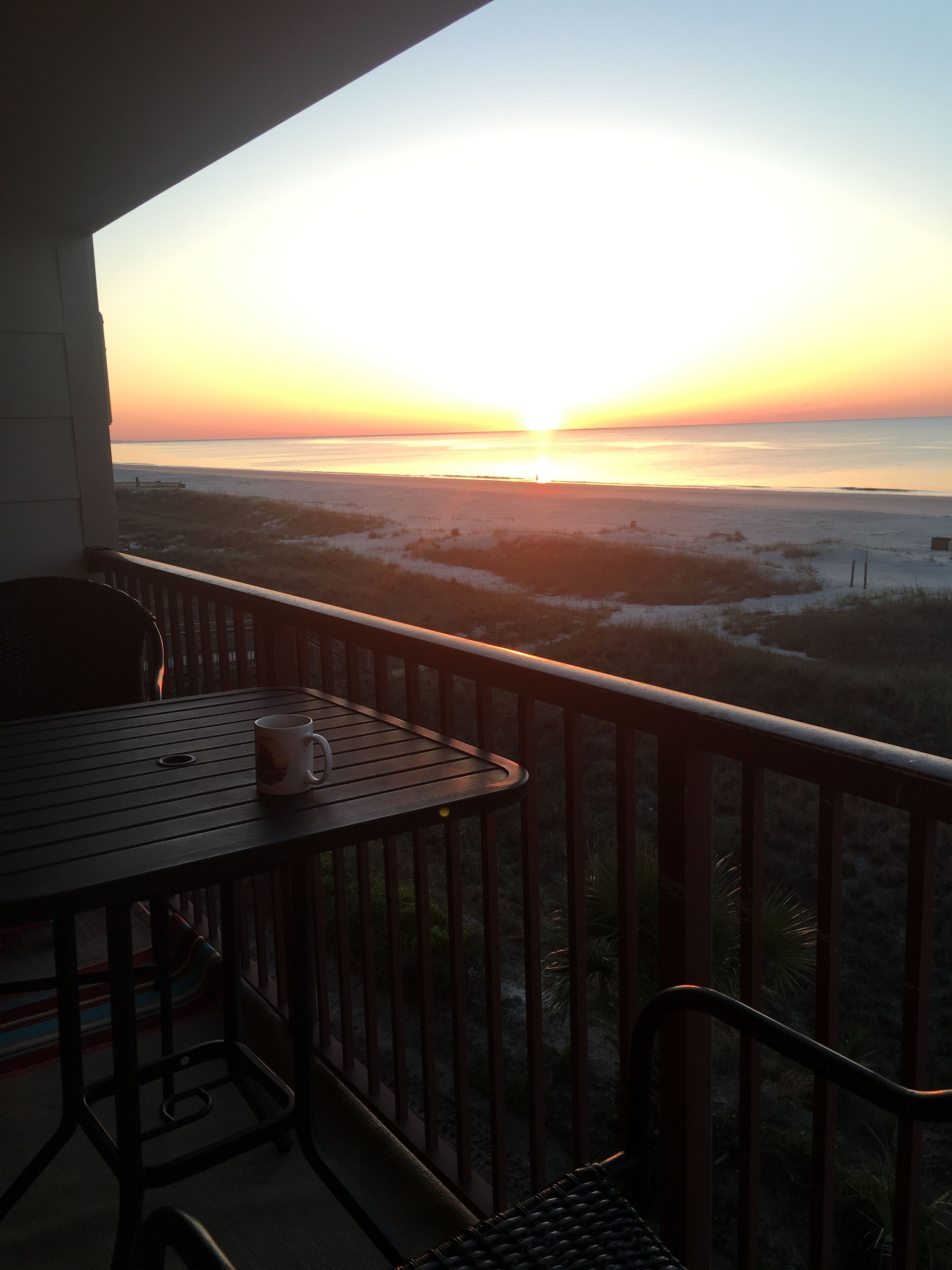 A balcony with a table and chairs, overlooking a beach at sunset.