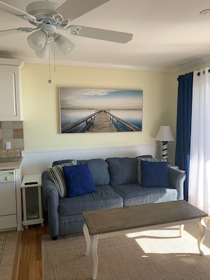 A living room with a grey sofa, a wooden coffee table, a ceiling fan, a painting of a pier, and a kitchen area with a white appliance. - Panoramic sea views in Admirals Row (Hilton Head Island)