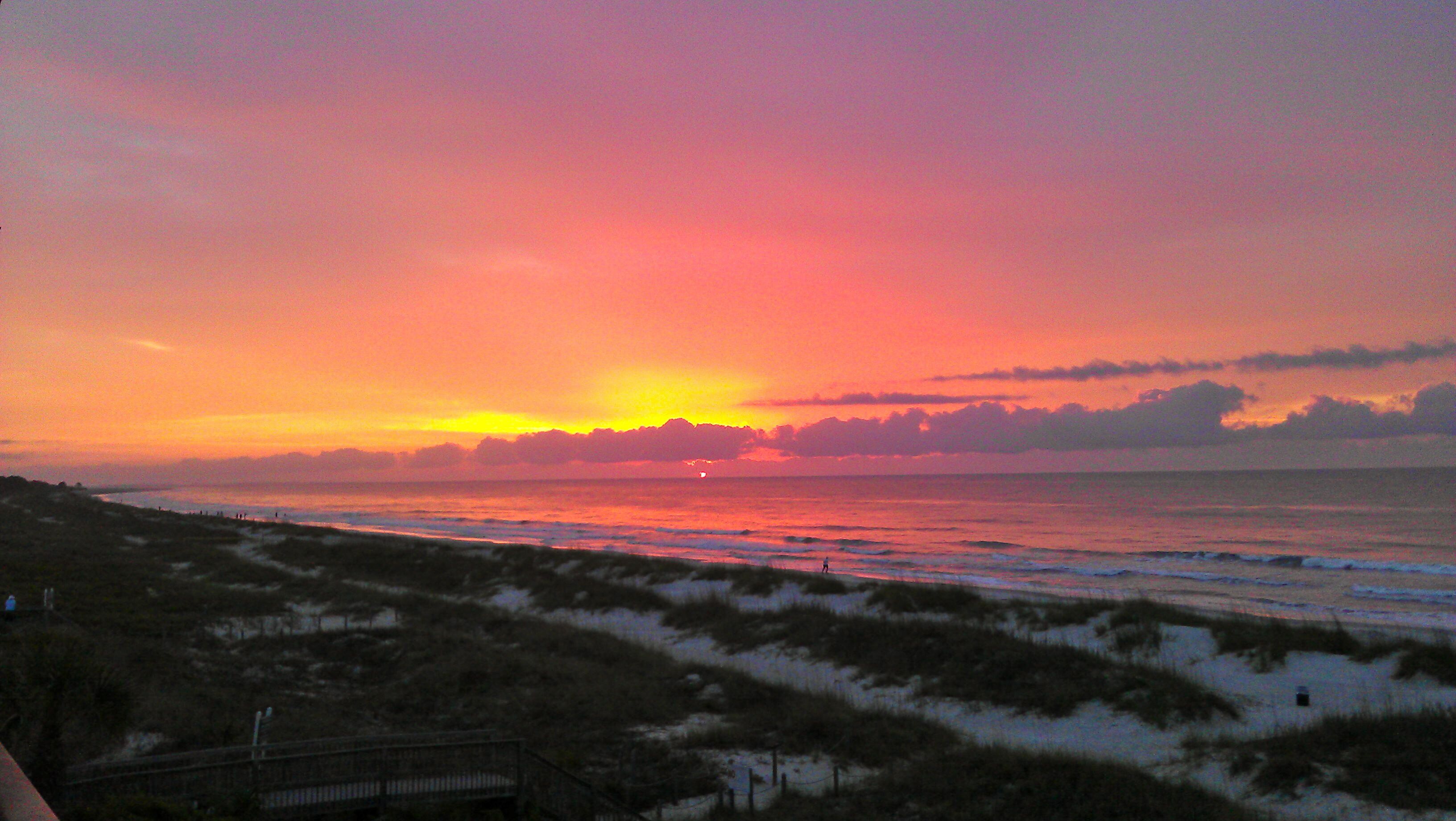 Ein Sonnenuntergang über einem Strand mit Dünen und einem Promenadenweg.