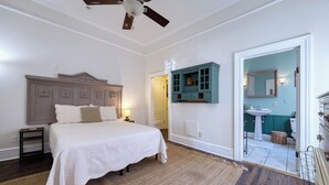 Une chambre à coucher comprenant un lit, une table de chevet, un ventilateur de plafond, un meuble fixé au mur et une salle de bain visible à travers une porte ouverte.