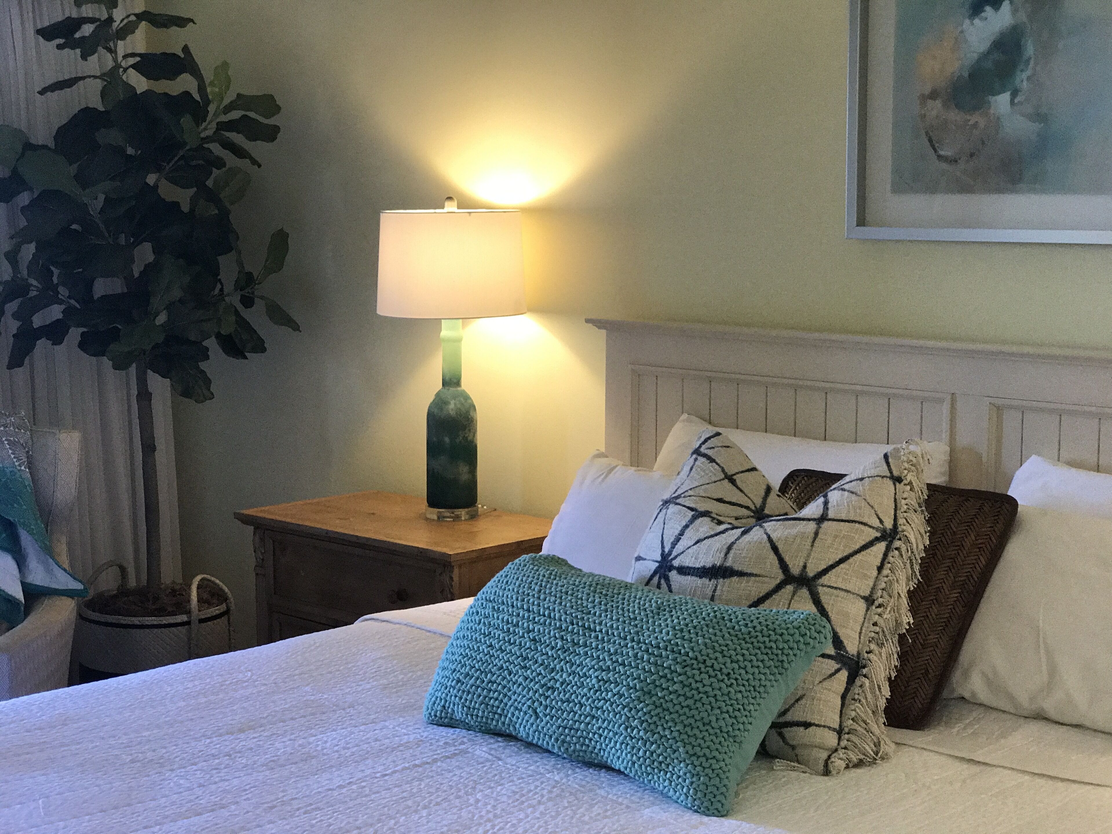 A neatly made bed with a white comforter and several pillows, including a green knitted one. A bedside lamp with a green shade is on a wooden nightstand beside the bed. A large green plant is visible in the corner.