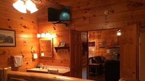 Interior de una cabaña de madera con un banco, un televisor montado en la pared, un baño con lavamanos y espejo, y un comedor con mesa y sillas.