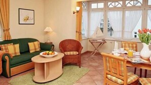 A living room with a green sofa, a round coffee table, a wicker chair, a dining table with chairs, and a window with curtains.