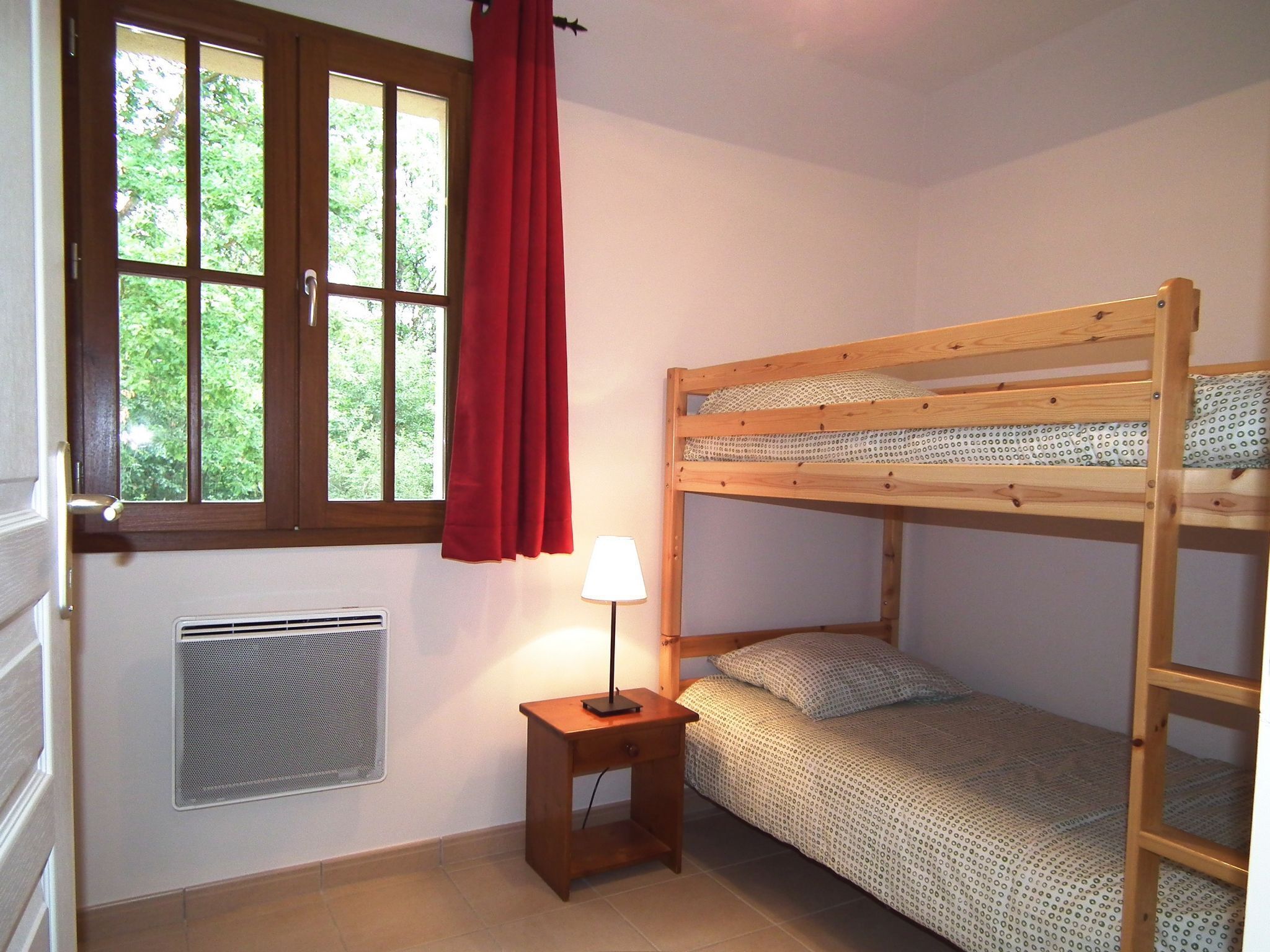 A small, tidy room with a bunk bed, a window with curtains, a heater, and a bedside table with a lamp.