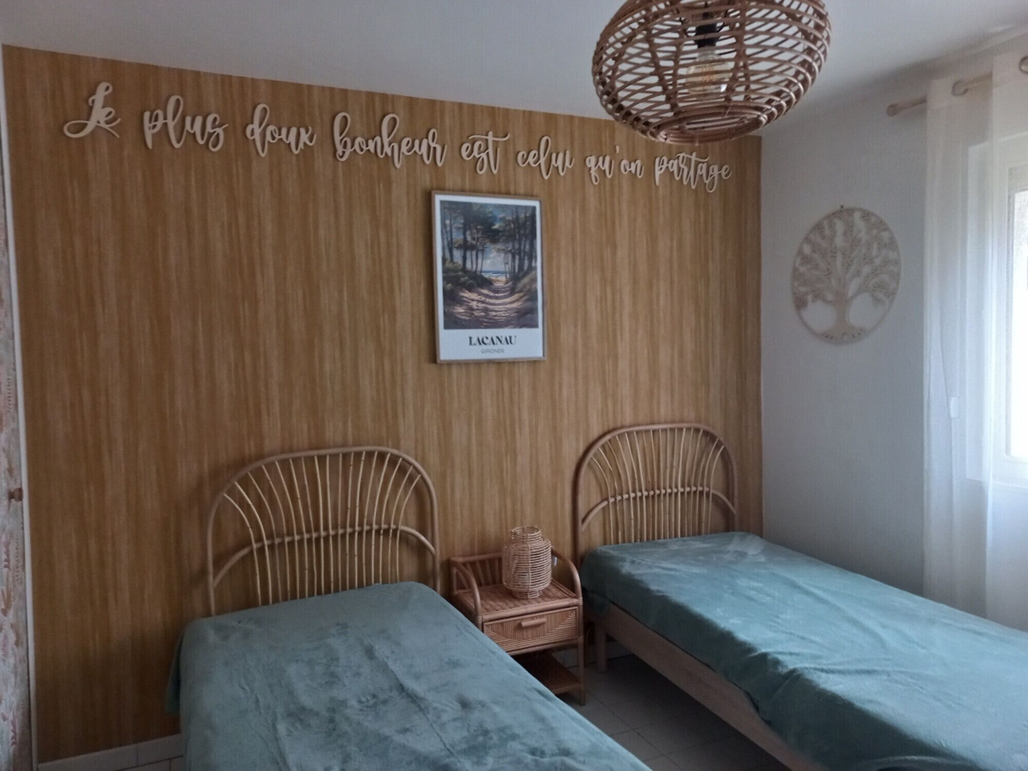 Un dormitorio con dos camas individuales, una silla de mimbre, un cuadro enmarcado y una pared con una frase en francés.