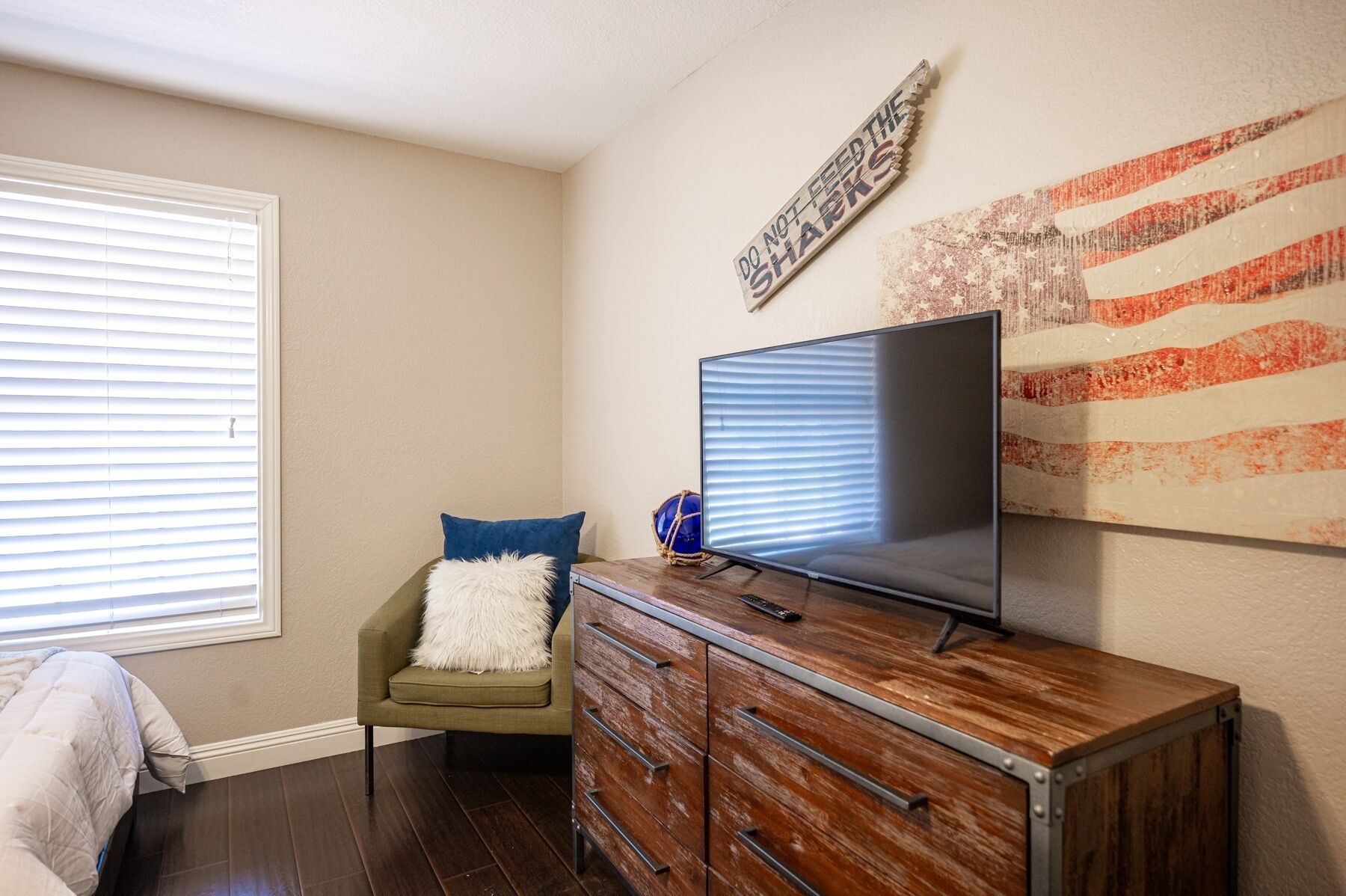 A living room with a flat-screen TV on a wooden cabinet, a green armchair with a white pillow, and a wall with a 'Do Not Feed the Sharks' sign.