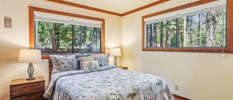 Un dormitorio con cama, dos mesitas de noche, dos lámparas y ventanas amplias con vistas al bosque.