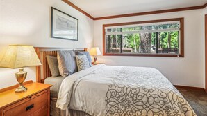 A bedroom with a large window, a bed with patterned bedding, a nightstand with a lamp, and a framed picture on the wall.