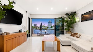 A modern living room with a wooden cabinet, a flat-screen TV, a coffee table, a white sofa, and a view of a cityscape through large glass doors.