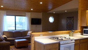 Uma sala de estar acolhedora com um sofá e um banquinho, uma cozinha com armários de madeira e um fogão, e uma área de jantar com uma mesa posta para dois.