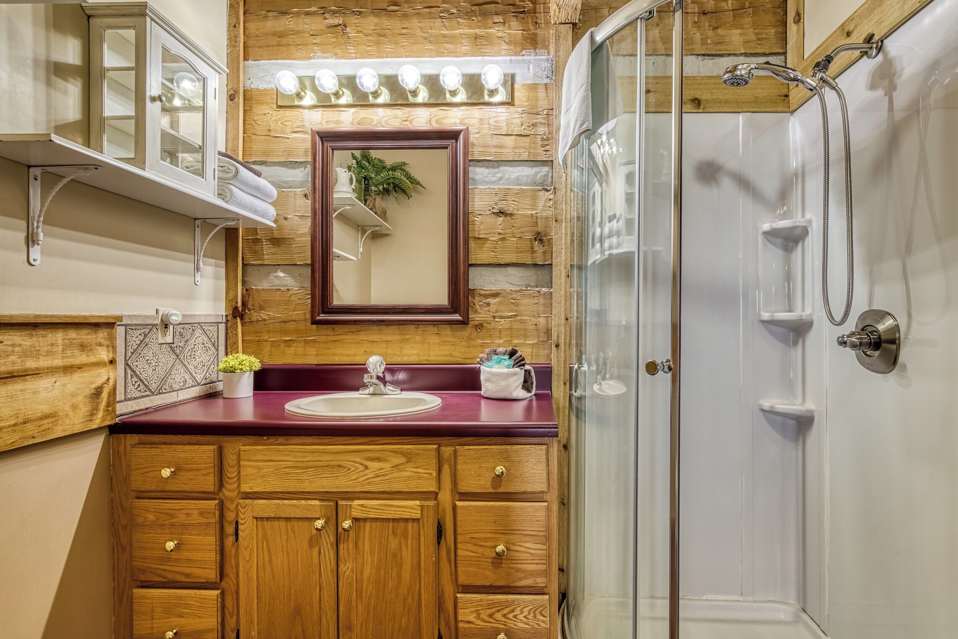 Un baño con muebles de madera, encimera de mármol, lavamanos, espejo y cabina de ducha con mampara de vidrio.