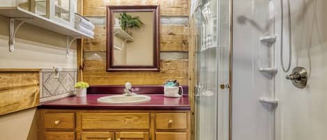 Un baño con muebles de madera, encimera de mármol, lavamanos, espejo y cabina de ducha con mampara de vidrio.