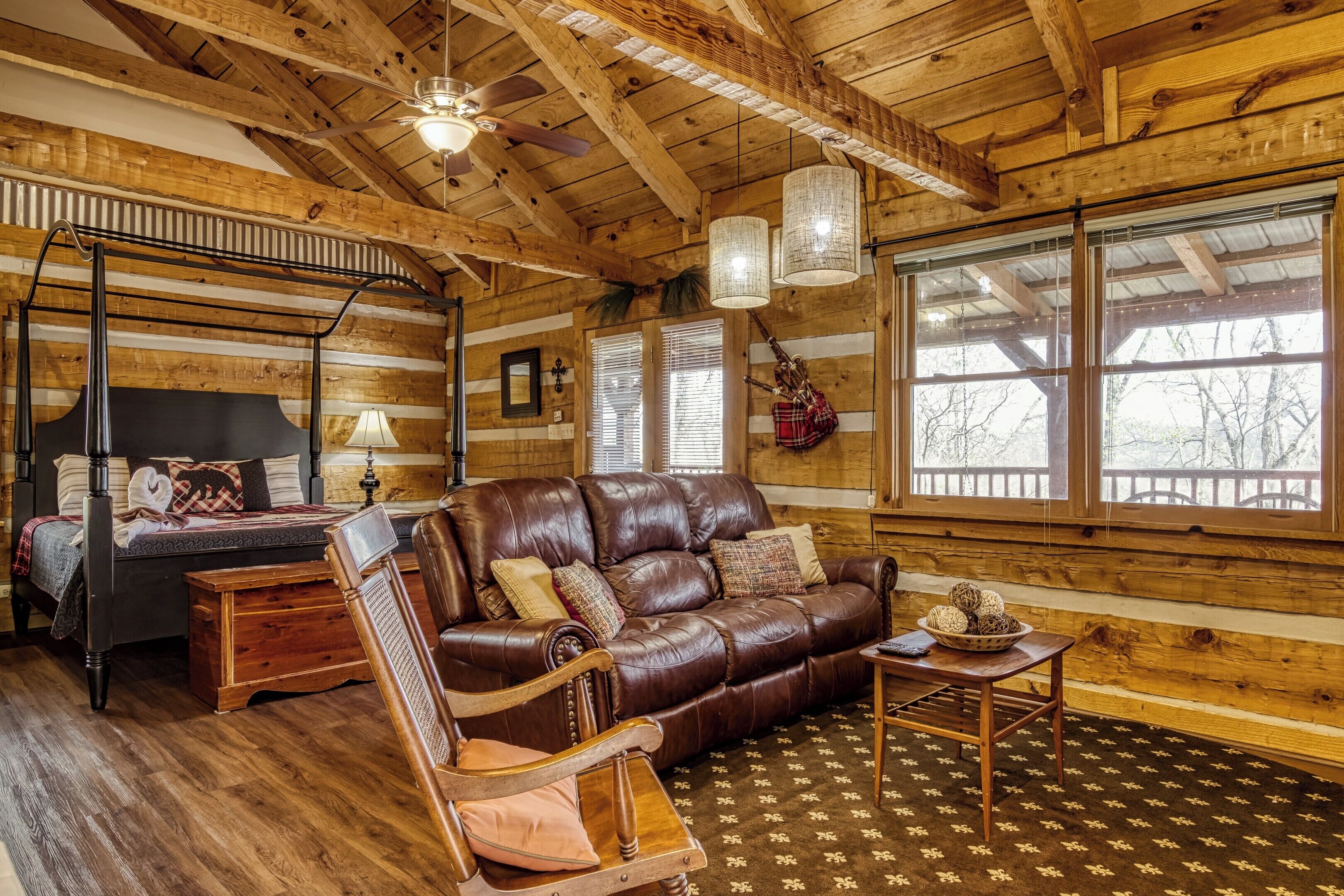 A log cabin interior with a bed, leather sofa, wooden rocking chair, and large windows.