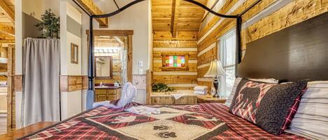 A room with a bed, wooden ceiling beams, a window with blinds, and a small table with a lamp.
