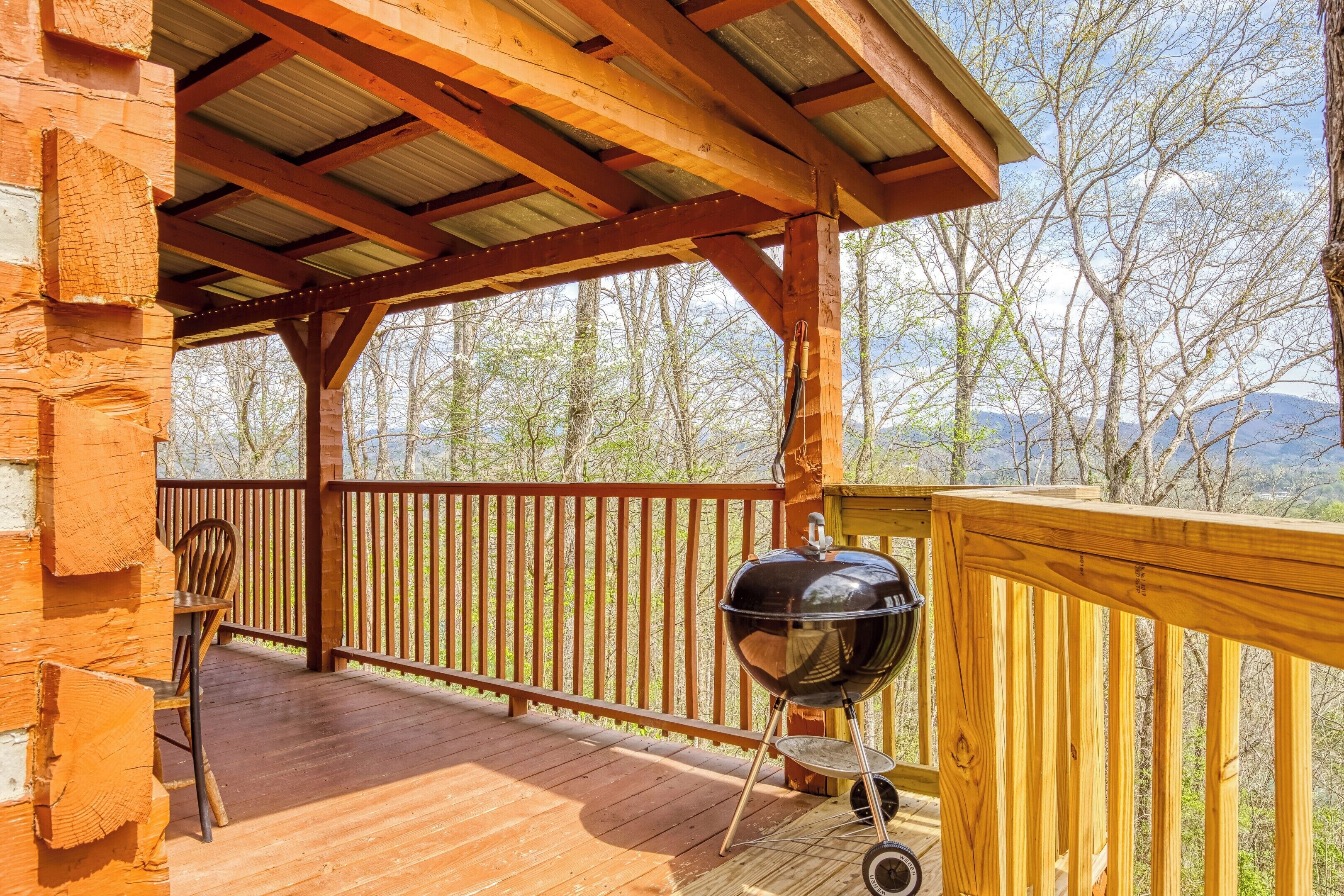 A covered deck with a grill, wooden railing, and a view of trees and mountains.