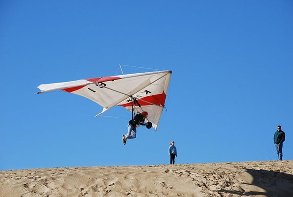 A paraglider in mid-flight over a desert landscape with two observers on the ground. - Semi oceanfront location with a 3 min walk to the ocean and Gift Card (Nags Head)
