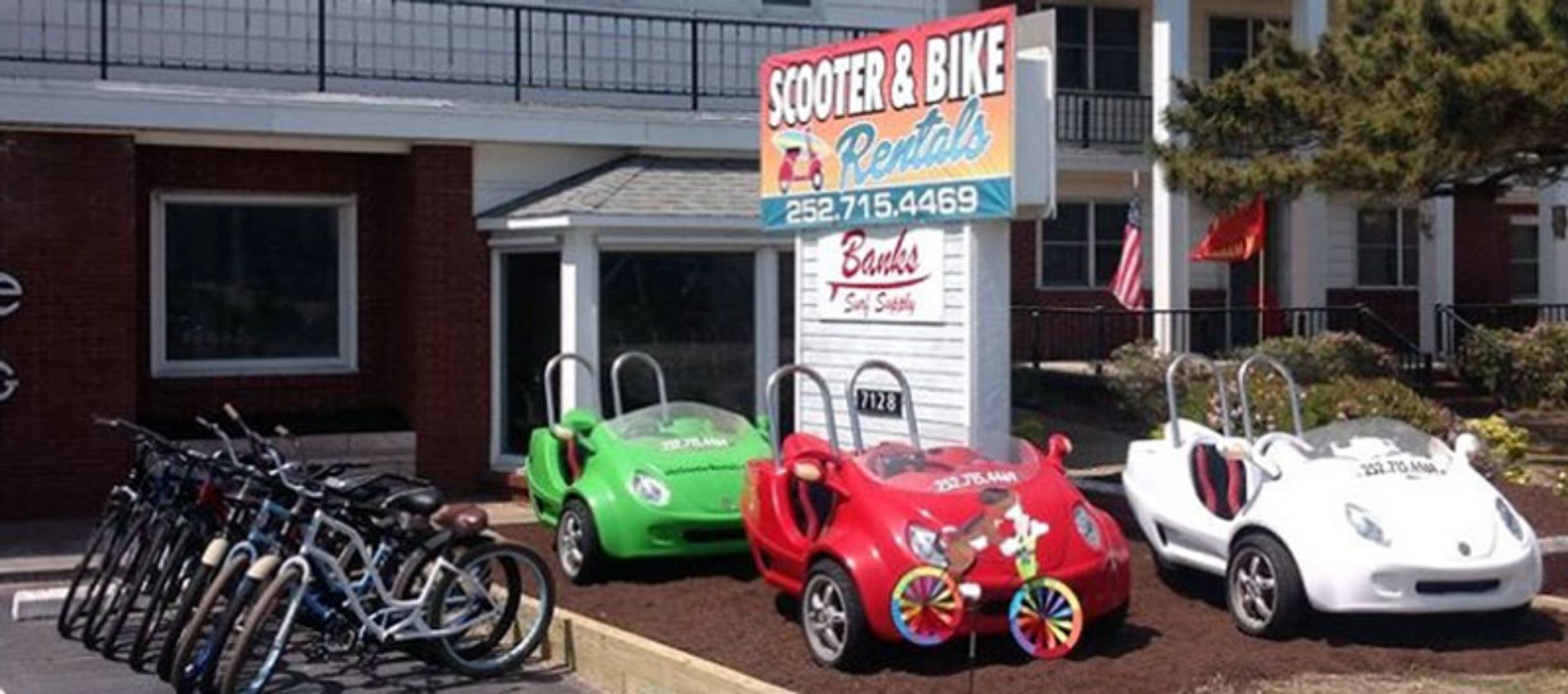 A building with a sign for scooter and bike rentals, a parking lot with bicycles, and a storefront with a sign for a bank.
