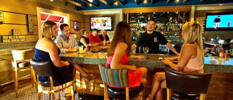 A bar area with a group of people seated at a counter, a bartender behind the bar, and a television screen displaying a sports event.
