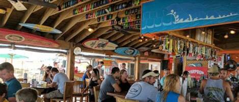 Um restaurante ao ar livre movimentado, com vigas de madeira e guarda-sóis coloridos.