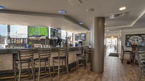 A bar area with wooden flooring, bar stools, a counter with a television displaying a sports game, and a view of the outdoors through large windows. - Free Beach SRVC! Direct Ocean Views! Free Beach Bonfires! Great Amenities! (Miramar Beach)