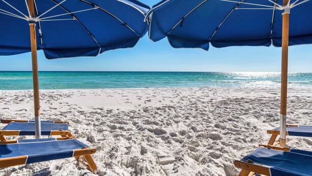 Deux parasols de plage bleus et des chaises longues en bois sur une plage de sable, avec une eau bleu clair en arrière-plan.