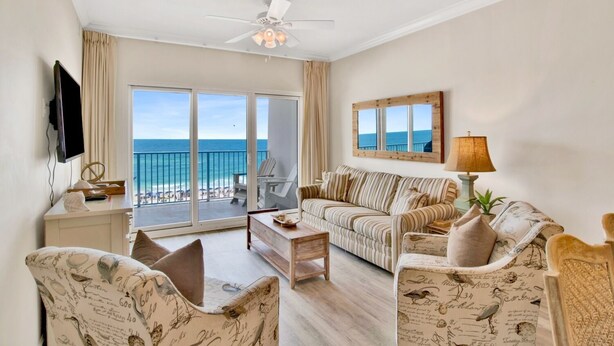 A spacious living room with a large sliding glass door leading to a balcony with a view of the ocean. There is a ceiling fan, a flat-screen TV mounted on the wall, and a sofa with striped cushions.