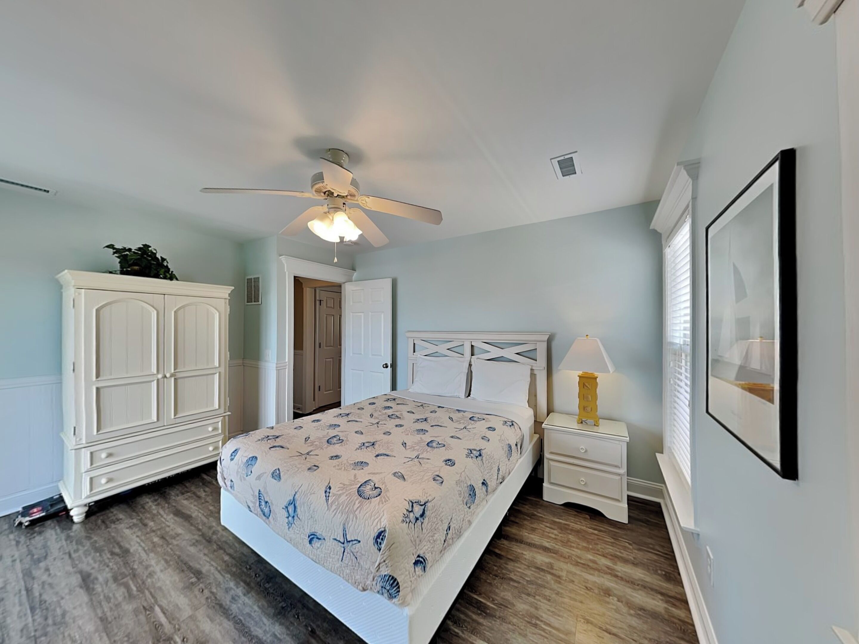 Une chambre à coucher comprenant un lit, une table de chevet, une commode, un ventilateur de plafond et une porte.