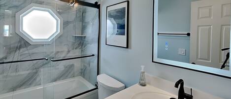 A modern bathroom with a glass shower enclosure, a white toilet, a sink with a black faucet, and a framed picture on the wall.