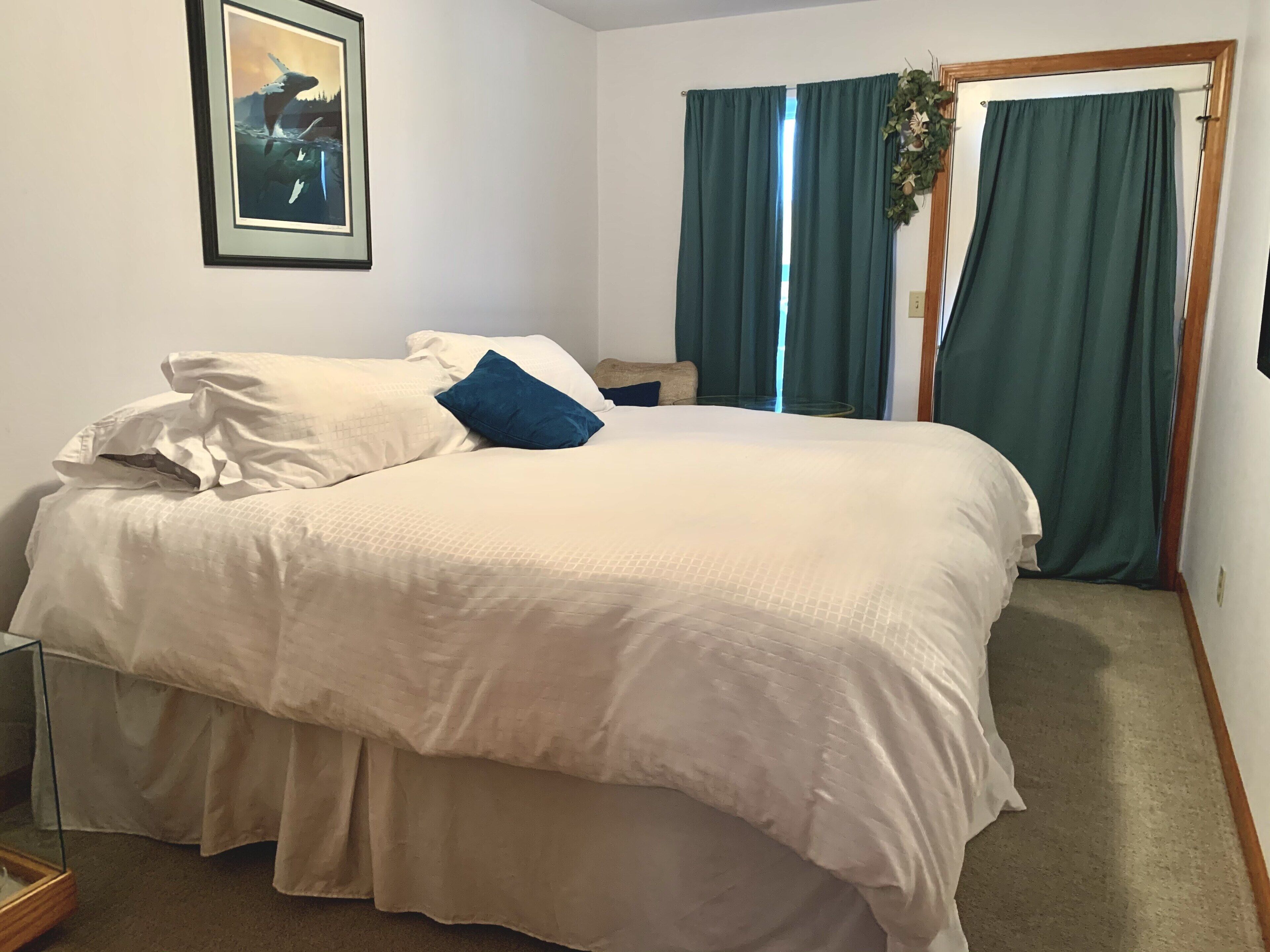 A neatly made bed with white and blue bedding, a framed picture on the wall, and a window with green curtains.