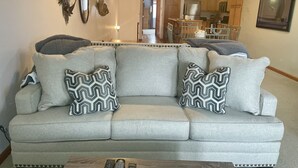 A living room with a grey sofa, patterned throw pillows, a coffee table, and a kitchen in the background.