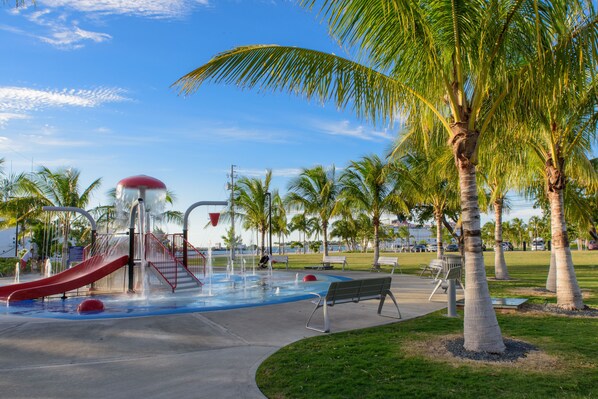 Explore the serene surroundings of this park, featuring a water play area with a slide and fountain, surrounded by palm trees and a paved walkway with benches.