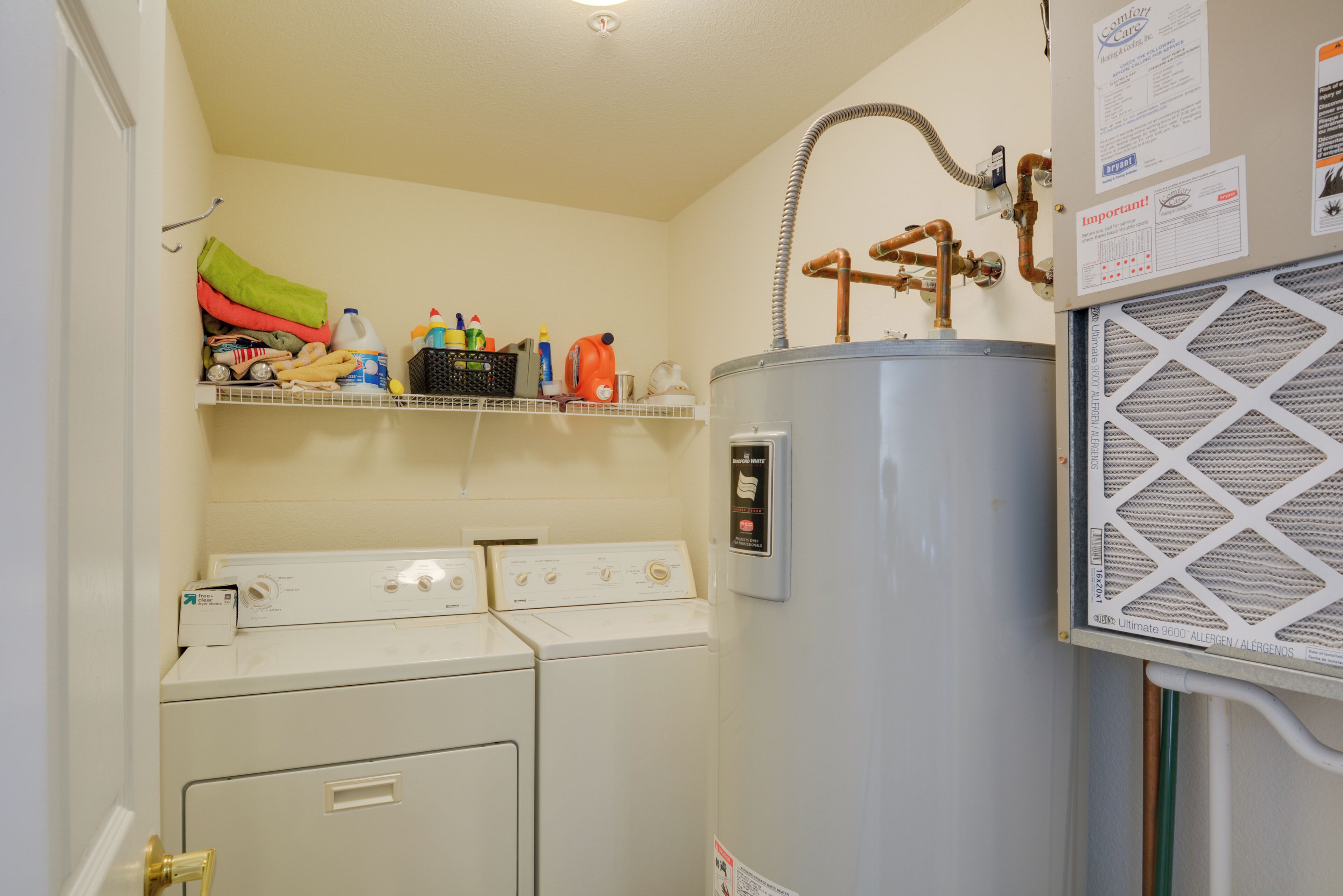 A laundry area with two washing machines, a water heater, and a shelf with various items.