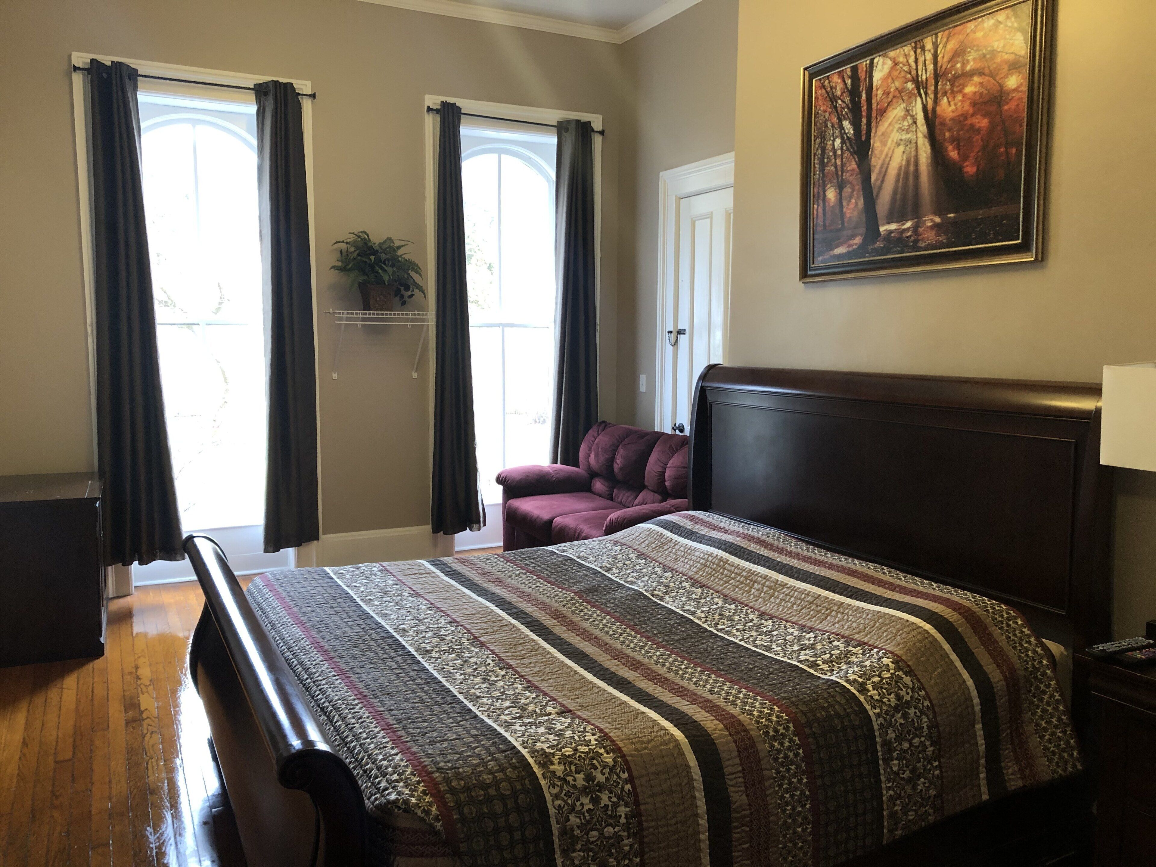 A bedroom with a large bed, a dark headboard, a window with curtains, a door, and a painting on the wall.