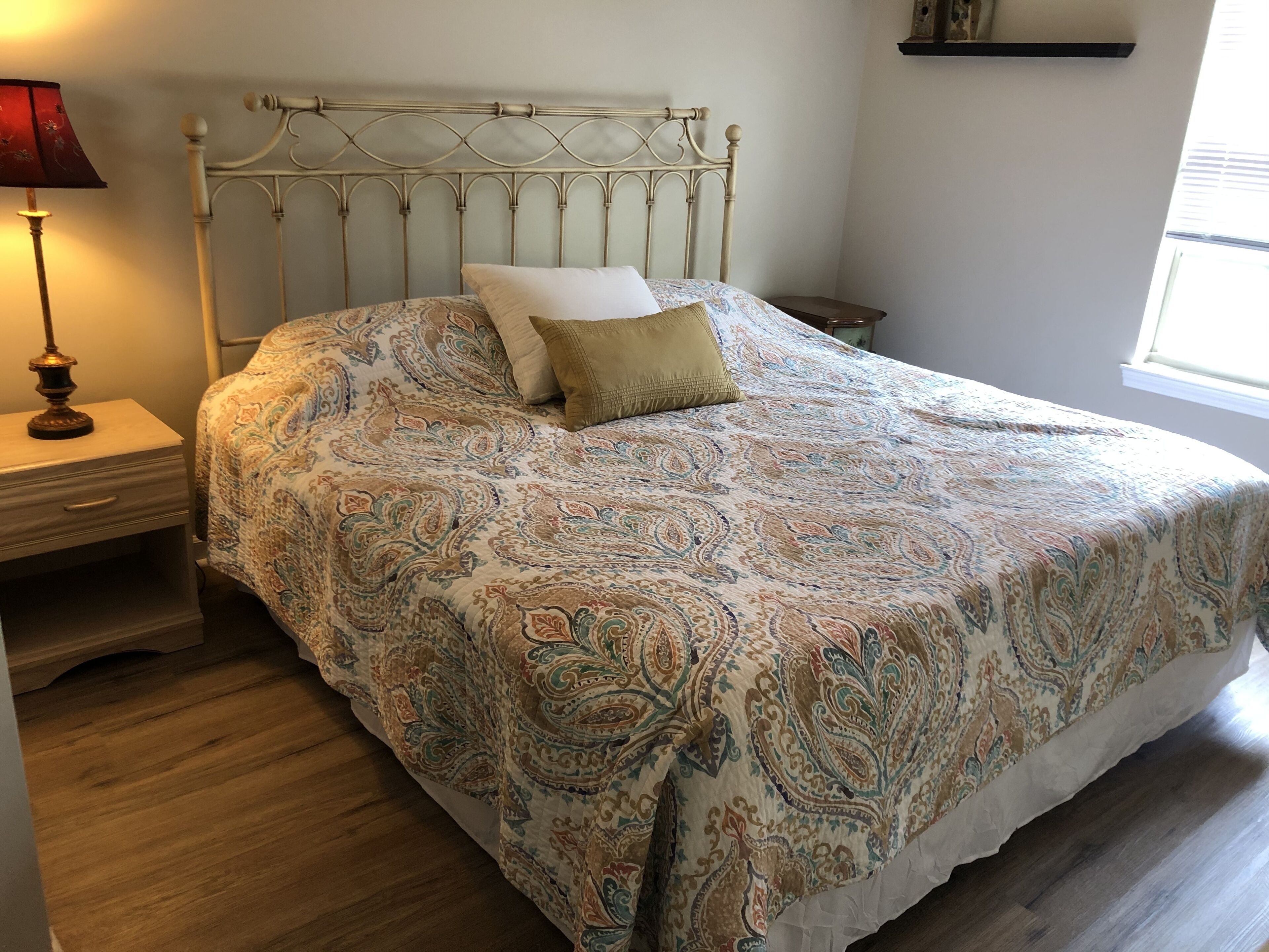 A bed with a patterned bedspread, a bedside table with a lamp, and a window with blinds.