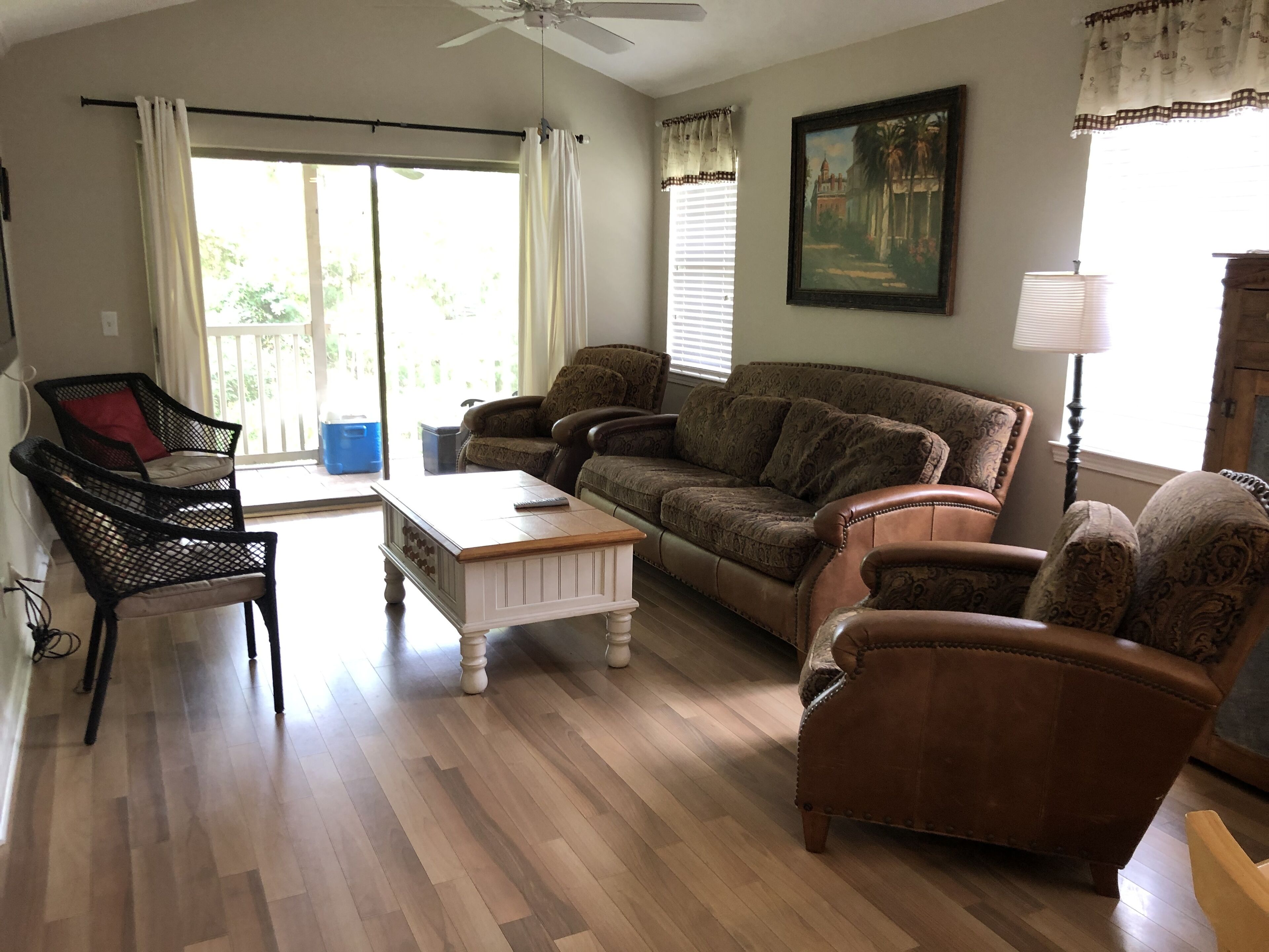 A living room with a brown sofa, a white coffee table, and a black chair. There is a painting on the wall and a ceiling fan.