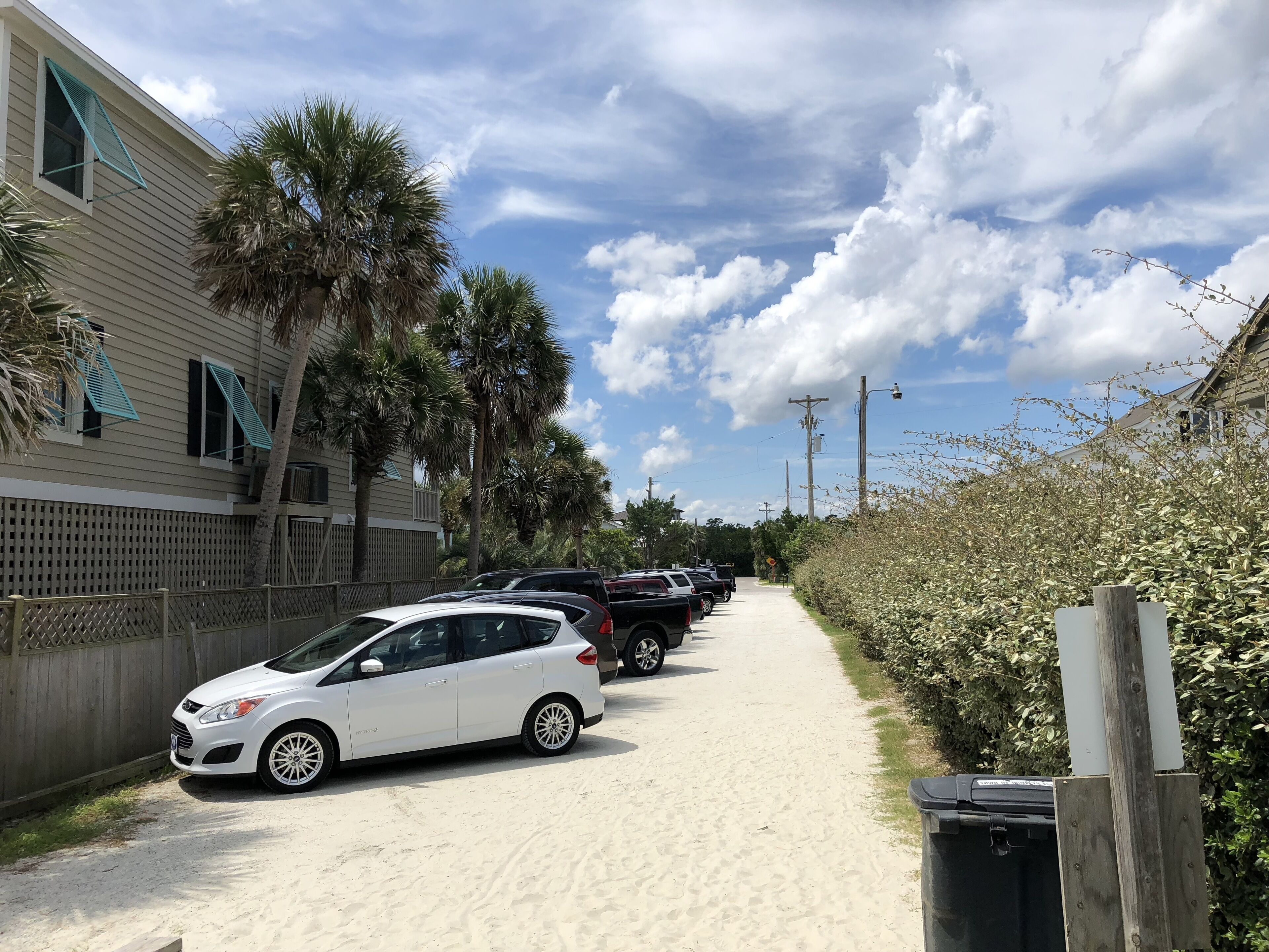 A pathway lined with parked cars and palm trees, leading to a building with a wooden fence and a beachside view.