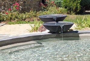 Una fuente con agua fluyendo hacia una pila, rodeada por un jardín con plantas y un muro de piedra.