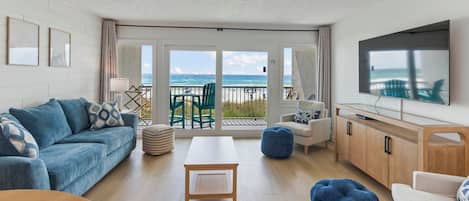 A spacious living room with a view of the ocean, a ceiling fan, a flat-screen TV, and a wooden dining table.