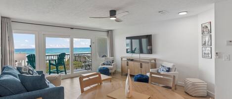 Un amplio salón con un gran ventanal con vista al océano, una mesa redonda de madera y un ventilador de techo.