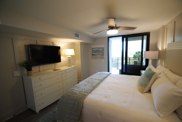 A bedroom with a bed, a TV mounted on the wall, a ceiling fan, and a window with a view.