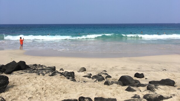 Een strand met heldere lucht, golven en een persoon in rood.