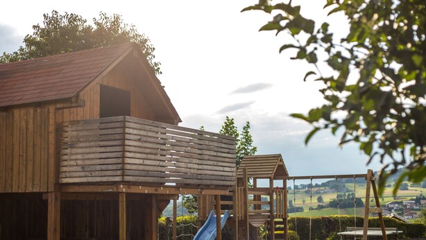 A wooden play structure with a slide and swings, set against a rural backdrop. - Gästehaus Weber (Arrach)