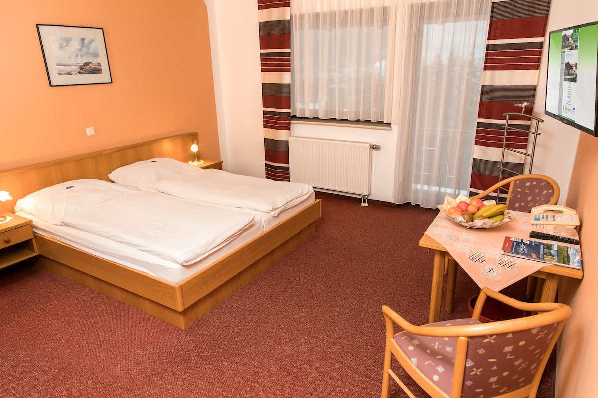 Habitación de hotel con cama doble, una mesita con frutero, una silla y un escritorio con computadora.