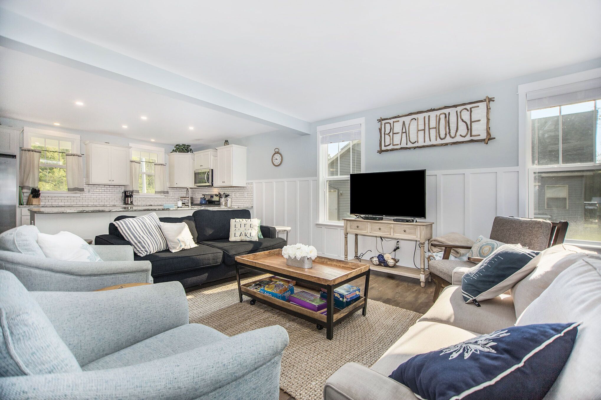 A modern living room with a kitchen in the background, a flat-screen TV, and a sign that reads "BEACHHOUSE".