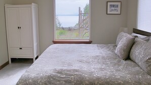 A bedroom with a large bed, a wardrobe, and a window with a view of a body of water and greenery. - Beautiful Oceanfront ‘Cottage’ - Breathtaking Views,Beach,Pet Friendly,Fireplace (Denman Island)