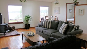 A living room with a sofa, a coffee table, and a fireplace. There are two windows and a potted plant. - Beautiful Oceanfront ‘Cottage’ - Breathtaking Views,Beach,Pet Friendly,Fireplace (Denman Island)