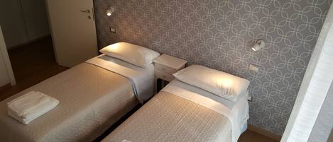 Une chambre d’hôtel avec deux lits simples, chacun muni d’un oreiller et d’une serviette pliée. Il y a une petite table de chevet avec une lampe sur chaque lit. Les murs sont ornés d’un motif géométrique.